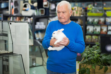 Senior European man standing in salesroom of pet shop and holding white rabbit in hands.