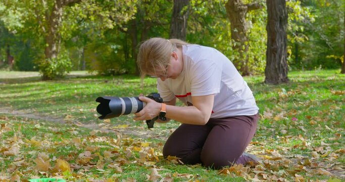 Young curvy blonde woman makes photos holding professional camera and sitting on knees on grass with yellow leaves IN autumn park