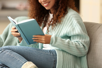 Obraz premium Young African-American woman in blue cardigan reading book on sofa at home