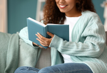 Fototapeta Young African-American woman in blue cardigan reading book on sofa at home obraz