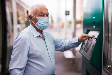 European old man in face mask standing on tram stop and using cash machine with credit card.