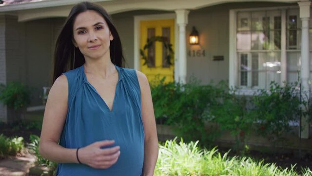 MCU portrait of a pregnant woman standing in front of suburban home looking into camera confidently.