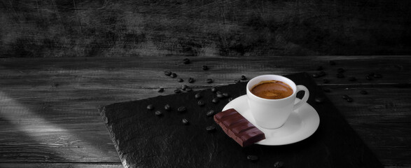 A white porcelain cup of coffee with tight crema and chocolate on a slate, dark wooden table, the ray of sunlight, scattered coffee beans, toned b&w
