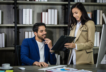 Millennial Indian Asian male successful executive businessman manager in formal suit holding clipboard discussing with female secretary businesswoman colleague at working desk in company office