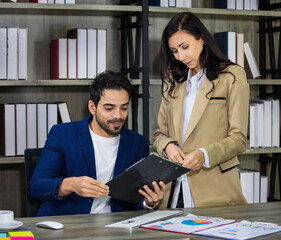 Millennial Indian Asian male successful executive businessman manager in formal suit holding clipboard discussing with female secretary businesswoman colleague at working desk in company office