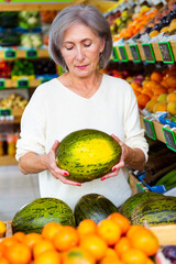 Woman chooses watermelon in greengrocer. Man walking in background