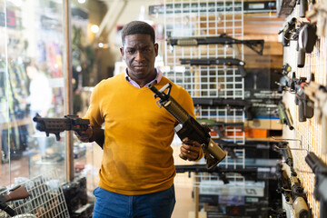African-american man choosing assault rifle and shotgun in airsoft shop.