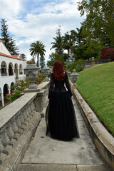 Fototapeta premium portrait of pretty female model with red hair wearing glamorous gothic black lace ballgown. Posing in a fairytale castle location with staircases