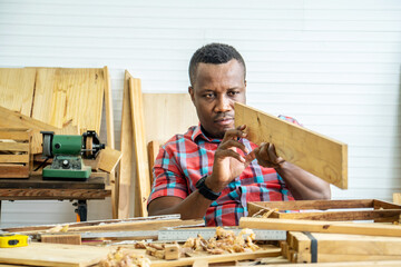 Young carpenter african american man looking and choosing wood and using sandpaper to rub wooden plank at workshop table in carpenter wood factory