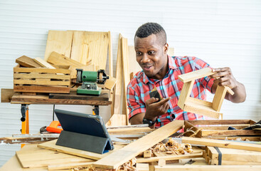 Young african american male carpenter sitting at table showing wooden items through tablet while video online chatting with customer or teaching online in factory