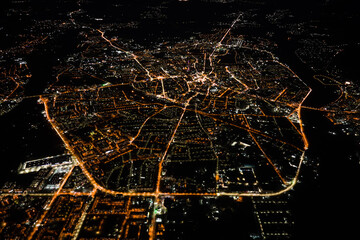 Aerial view from airplane window of buildings and bright illuminated streets in city residential area at night. Dark urban landscape at high altitude