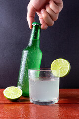 Lime drink in a glass and hand holding glass bottle over red wooden table.