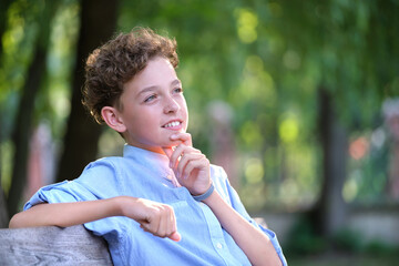 Young happy child boy relaxing sitting on bench in summer park. Positive kid enjoying summertime outdoors. Child wellbeing concept