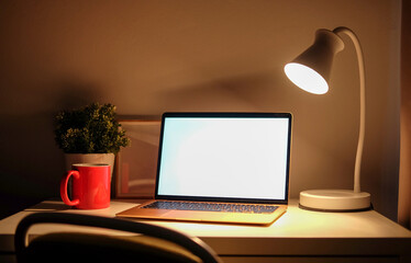 Computer laptop with blank screen, coffee cup and lamp on white table in dark room.