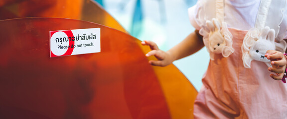 Portrait child's hand touching an orange acrylic sheet that is labeled in Thai and English: Please do not touch.