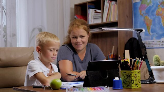 A small schoolboy boy and his mother are sitting at a desk and talking to a teacher via video link while doing homework on distance learning. The concept of the coronavirus.