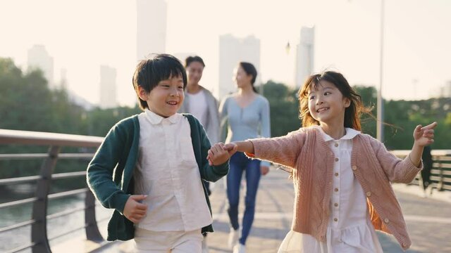 two happy asian children walking in park with parents in background