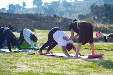 Senior woman doing downward facing dog while instructor helps her, outdoor yoga group practice, exercise on mountain esplanade. Healthy, health, selective focus, blur