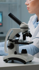 Close up of microscope tool while scientist analyzing petri dish and using modern computer in background. Woman at laboratory desk with blood sample and tray of vacutainers for examination