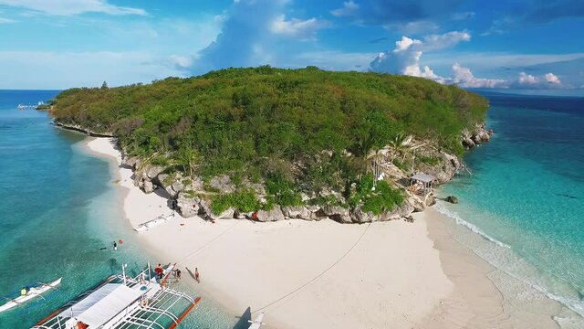 drone pull back view of a rocky island with lush green forests and boats on a white sand beach zooming out to reveal the blue green seabed of an island called Sumilon