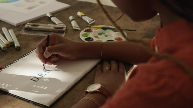 Creative young woman drawing the word ''DESIGN'' on a notepad. Female graphic designer planning a new design project while sitting in her home office.
