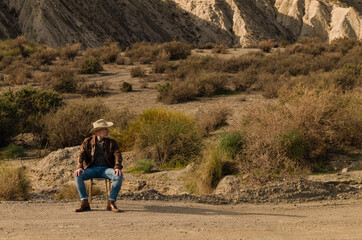 Adult man in cowboy hat sitting on abandoned chair in desert, Almeria, Spain