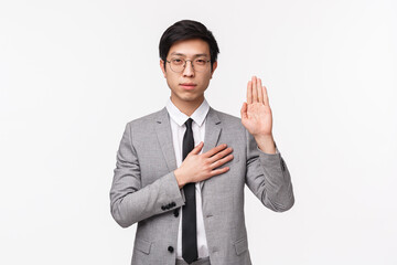 Waist-up portrait of devoted good-looking asian young man in grey suit, making promise, pledge or give oath, raise one hand and put palm on hear as being honest and sincere, white background - obrazy, fototapety, plakaty