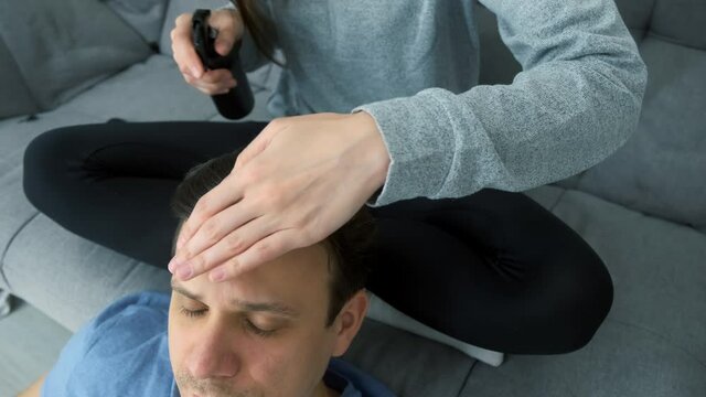 Woman is doing a man's hair while sitting at home on the couch. She sprays his hair with a styler and pins it with hairpins preparing for braiding pigtails. Evening of a young married couple together.