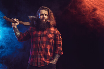 Naklejka premium Handsome man holds old axe. Bearded lumberjack. Serious brutal man in checkered shirt with long hair