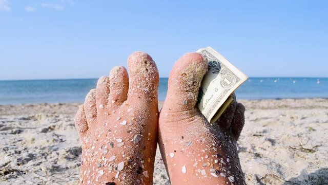 Two bare feet of caucasian woman soiled in sand with shell rock and dollar bill sandwiched between your fingers on sandy sea shore on sunny summer day. Concept of rest, relax,vacation, travel tourism