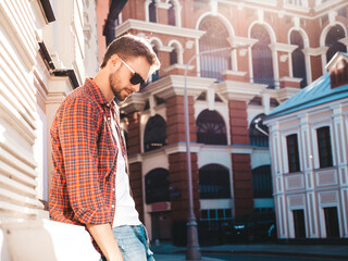 Handsome smiling stylish hipster lambersexual model.Modern man dressed in red checkered shirt. Fashion male posing near skyscraper on the street background in sunglasses. Outdoors at sunset