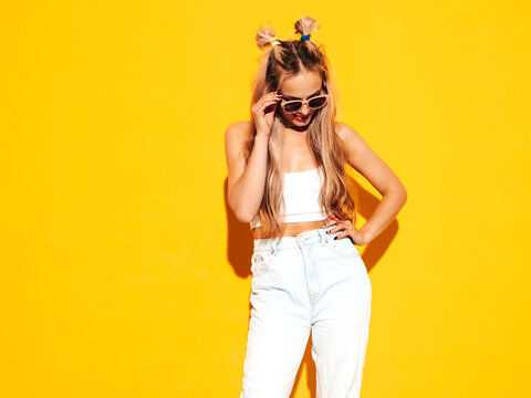 Portrait of young beautiful smiling blond female in trendy summer clothes. Sexy carefree woman posing near yellow wall in studio. Positive model having fun indoors. Cheerful and happy in sunglasses