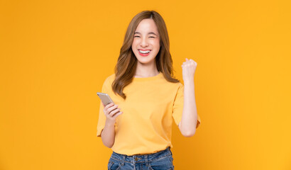 Cheerful beautiful Asian woman holding smartphone with fists clenched celebrating victory expressing success on light orange background.