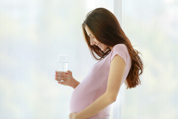 Caucasian young healthy female big belly tummy prenatal pregnant mother in casual shirt standing in front of curtain background smiling holding glass of drinking water at home in morning alone at home