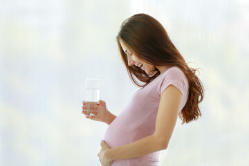 Caucasian young healthy female big belly tummy prenatal pregnant mother in casual shirt standing in front of curtain background smiling holding glass of drinking water at home in morning alone at home