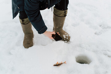 Fisherman photographing caught fish lying on ice with smartphone camera. Winter fishing