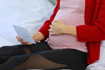 Obraz premium Caucasian millennial young happy healthy female prenatal pregnant mother in casual outfit sitting smiling on bed in bedroom showing ultrasound Xray picture to family via video call with smartphone