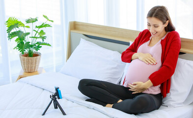 Caucasian millennial young happy healthy female prenatal pregnant mother model in casual outfit sitting smiling on bed in bedroom holding hands on big belly with love using smartphone video call.