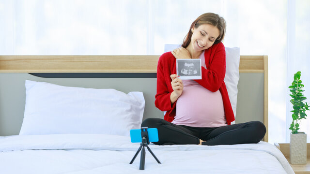 Caucasian millennial young happy healthy female prenatal pregnant mother in casual outfit sitting smiling on bed in bedroom showing ultrasound Xray picture to family via video call with smartphone