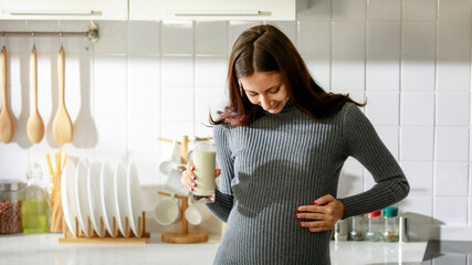 Caucasian millennial young happy female prenatal pregnant mother in casual outfit standing alone holding hand on big belly tummy with love drinking fresh milk for breakfast in full equipment kitchen