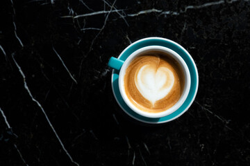 Cup of coffee latte in yellow ceramic mug, milk froth, heart shape on black table, fresh breakfast.