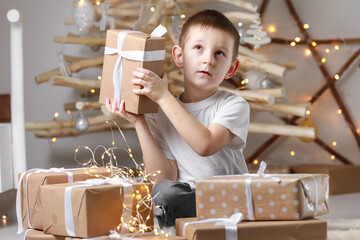Holidays Concept. A smiling little excited boy child is sitting by the wooden decorations Christmas tree and many gift boxes, holding a gift box. new Year's Eve and Christmas, waiting for a miracle