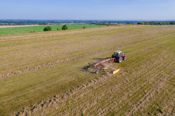 Aerial view on a tractor that collects hay in rows with a disc rake. Farmer stores fodder for cattle in summer season.