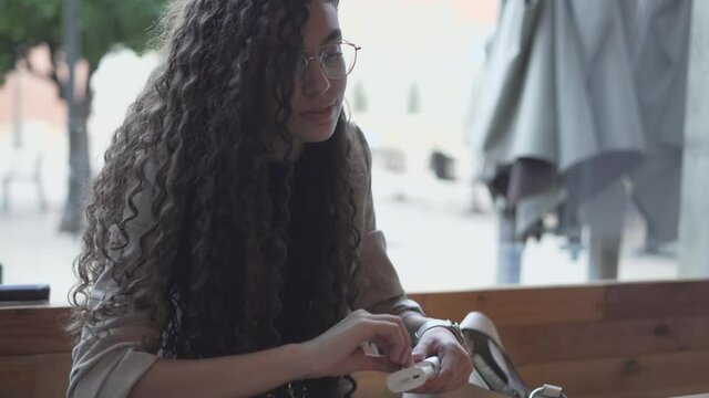 Beautiful Young Woman In Eyeglasses Puts On Bluetooth Earbuds While Sitting Outdoor. medium shot
