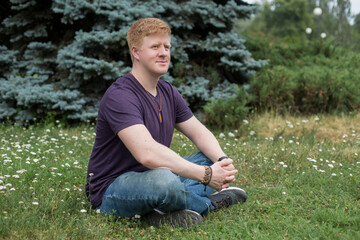 Naklejka na ściany i meble Portrait of redhead caucasian man wearing purple t-shirt and blue jeans and sitting on the grass in city park during lunch break at summer. Person looks away. Theme of rest in work break.