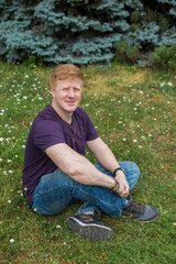 Naklejka na ściany i meble Portrait of redhead man wearing purple t-shirt and blue jeans and sitting on the grass in city park during lunch break at summer. Person looks at camera and smiles. Theme of rest in work break.