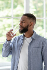 Millennial african american guy with beard and perfect skin drinks clean water from glass with closed eyes