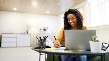 Fototapeta Black woman sitting at desk, using pc writing in notebook obraz