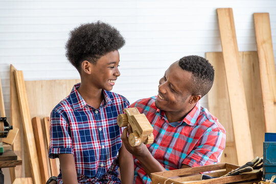 Happy African Black Male Carpenter Man Father And Boy Kid Child Son With Wooden Toy Car Model Present Gift In Carpentry Workshop. Concept Hobby At Home.