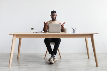 Handsome millennial black man working on laptop, making video call, having remote job, sitting at desk in home office